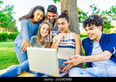 I giovani amici hipster si siedono sull'erba nel parco della città, creando volti a sorpresa che guardano lo schermo del computer portatile. Concetto di divertimento moderno con milleniali su nuova tendenza Foto Stock