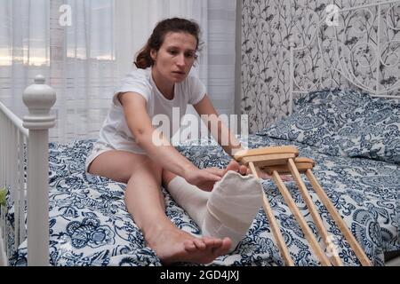 Una donna massaggia le dita di una gamba rotta con le mani. Vista dall'alto di una gamba rotta in un calco di gesso e stampelle. Riabilitazione domestica dopo un rotto Foto Stock