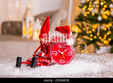 Elfo in feltro decorativo, sveglia rossa vintage con cappello di Natale su neve artificiale, albero di Natale offuscato con regali e bianco illuminato. Foto Stock