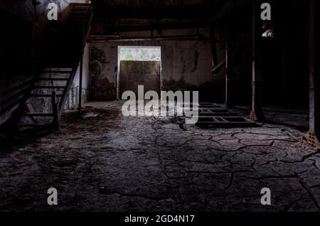 Vecchio sfondo interno di fabbrica. Camera oscura con sporcizia incrinata sul pavimento, scale di legno rotte e resti arrugginiti di macchinari Foto Stock