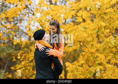 Una giovane coppia in un parco autunnale. Un uomo sorridente tiene una donna tra le braccia contro uno sfondo autunno colorato. Felice coppia sullo sfondo di autu Foto Stock