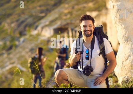 Ritratto di giovane uomo con zaino e fotocamera in montagna. Turisti felici e escursionisti offuscati in tour a piedi all'aperto Foto Stock