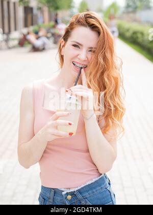 Giovane bella donna dai capelli rossi con bretelle bevande rinfrescare limonata all'aperto in estate. Ritratto di una ragazza sorridente con frettole. Foto Stock