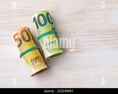 Rotolo di banconote da 100 euro e borra di moneta cartacea da 50 euro su una scrivania di legno bianco. Concetti di risparmio in contanti, conteggio di denaro, ricchezza e spese. Foto Stock