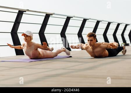 Attraente giovane coppia di fitness facendo esercizi di stretching all'aperto sul mare sul ponte, uomo sportivo e donna che si allenano insieme Foto Stock