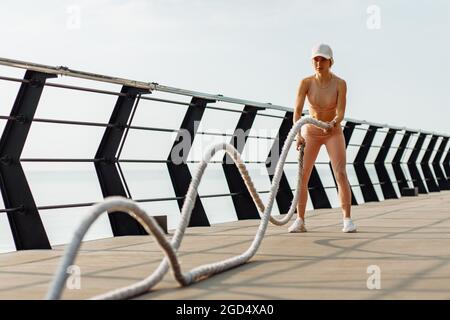 Fitness donna che si esercita con corde da combattimento all'aperto sul molo, donna in abbigliamento sportivo che fa allenamento corde da combattimento, braccia e cardio per exerci cross-fit Foto Stock
