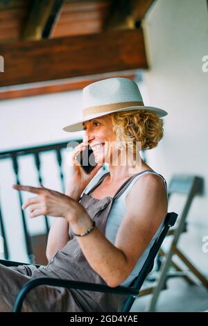 Donna bionda caucasica matura negli anni '50 che parla su un telefono cellulare sorridendo e indossando un cappello e seduto in una posizione rilassata. Concetto di stile di vita. Foto Stock