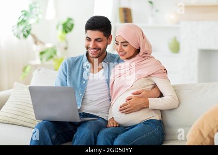 Amorevole coppia musulmana incinta scegliere mobili per la stanza del bambino in linea, usando il computer portatile mentre si riposa sul divano in soggiorno, spazio vuoto. Aspettatevi Foto Stock