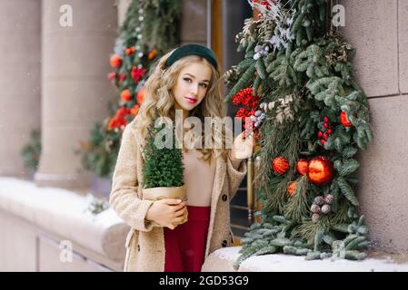 Una giovane donna hipster che cammina per le strade innevate della città tiene in mano un albero di Natale in vaso. In una gonna di borgogna e un cappotto beige Foto Stock