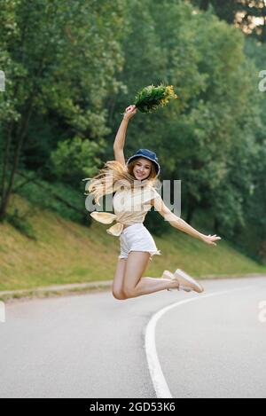 Una ragazza felice in pantaloncini bianchi e una blusa gialla salta con un bouquet di fiori, in piedi sulla strada vicino alla foresta Foto Stock