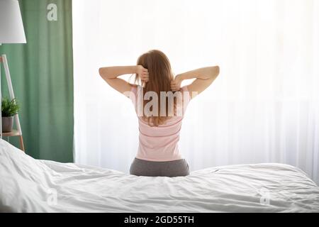 Vista posteriore della millenaria donna caucasica seduta sul letto dopo essersi svegliata a casa. Giovane donna che si è rilassato fine settimana mattina, stretching, sensazione di sonnolenza, Foto Stock