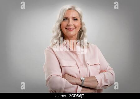 Studio ritratto di donna anziana con capelli grigi in piedi con braccia incrociate su sfondo grigio isolato. Spazio di copia. Foto Stock