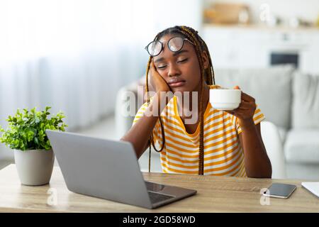 Donna nera esausta e millenaria freelancer seduto davanti al computer portatile con una tazza di caffè, sentendosi sonnolenta e stanca o sbornia, che soffre di burnou Foto Stock