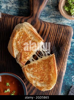 Vista dall'alto di un panino di formaggio alla griglia fatto in casa su un pannello di legno, servito con zuppa di pomodoro. Foto Stock