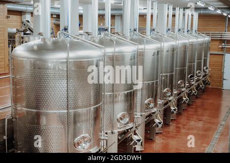 Fila di vasche di metallo per la maturazione del vino situata in sala illuminata di cantina contemporanea Foto Stock