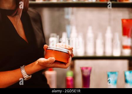 Foto ritagliata del parrucchiere femminile che tiene il vaso di maschera per capelli, donna coiffeur mostrando speciale prodotto di styling si leva contro sfondo sfocato della modalità Foto Stock