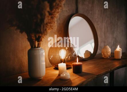 Specchio rotondo su mensola di legno con due candele brucianti e piccola testa di Buddha di porcellana riflessa in un angolo di superficie specchiata sopra la parete di cemento Foto Stock