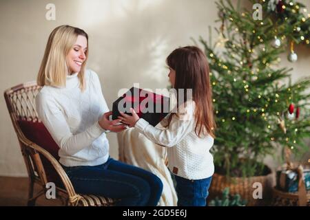 Figlia ragazza carina che dà il regalo di Natale la sua giovane madre sorridente, seduta in camera con albero decorato, mamma felice di famiglia e bambino che indossa maglia bianca Foto Stock
