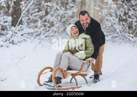 Felice giocosa coppia di famiglia matura che slitta nel parco invernale, ridendo e divertendosi insieme, positiva donna di mezza età seduta su slitta mentre il suo hus Foto Stock