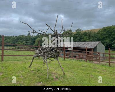 Il legno di spazzolone raccolto in campagna e trasformato in un modello fatto a mano di un cervo di Stag di fronte a stalle in un campo su una fattoria in Devon rurale Foto Stock