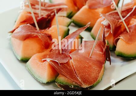 Diversi pezzi di melone con Prosciutto e stuzzicadenti in legno, disposti su un piatto bianco con fuoco selettivo in primo piano Foto Stock