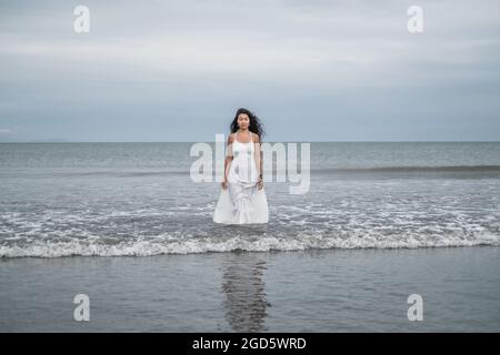 Affascinante giovane donna asiatica in abito bianco che esce dal mare. Capelli neri lunghi ricci. Foto romantica. Vista sull'oceano. Foto Stock