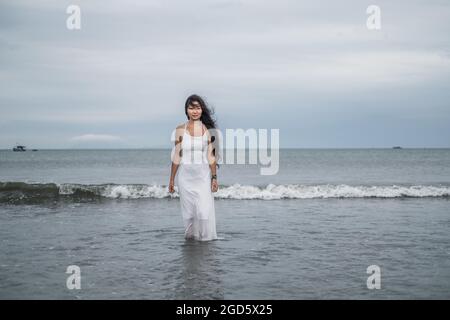 donna in abito bianco in piedi sulla spiaggia. Capelli neri lunghi ricci. Guardando la fotocamera con spazio per la copia. Foto romantica. Vista sull'oceano. Foto Stock