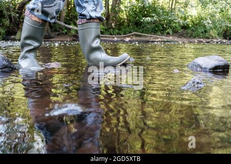 I piedi maschi in stivali impermeabili in gomma camminano lungo il fiume, sullo sfondo del verde, in una giornata estiva. Foto Stock