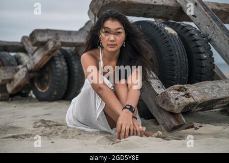 Giovane donna mongol di charme in abito bianco seduta sulla spiaggia e guardando la macchina fotografica con spazio di copia. Brunette capelli lunghi ricci. Foto romantica. Foto Stock