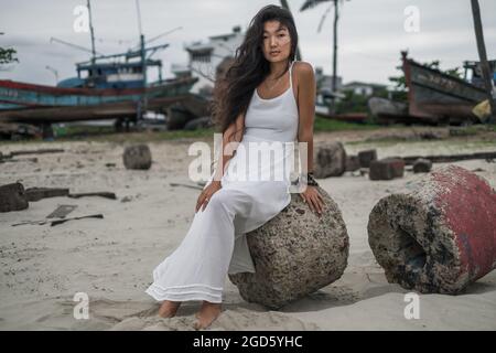 Bella giovane donna asiatica in abito bianco seduta sulla pietra e guardando la macchina fotografica con spazio di copia. Brunette capelli lunghi ricci. Romantico. Foto Stock