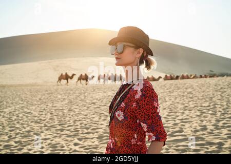 donna asiatica in abito rosso che guarda la vista nel deserto con caravan di cammelli e enormi dune di sabbia sullo sfondo Foto Stock