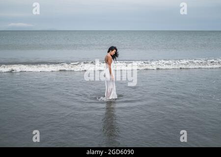 Giovane donna asiatica di charme in abito bianco che cammina in acqua nel mare. Capelli neri lunghi ricci. Guardando da parte. Foto romantica. Vista sull'oceano. Foto Stock