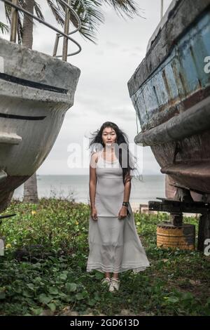 Affascinante giovane donna mongolo in abito grigio brillante in piedi tra due vecchie navi di legno. Capelli ricci. Foto romantica. Giornata ventosa. Foto di alta qualità Foto Stock