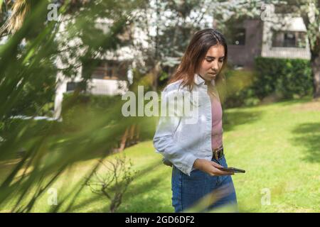 Ritratto attraverso il verde di una ragazza teenage Latina con il suo cellulare in mano come lei toglie la giacca in un parco Foto Stock