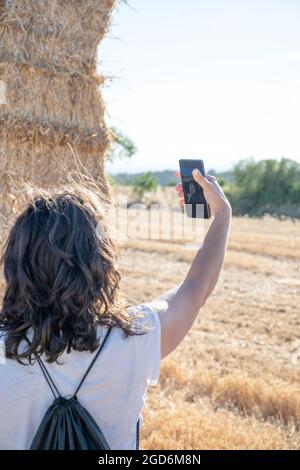 donna latina con bruna di mezza età che indossa una t-shirt bianca prendendo un selfie davanti ad alcune balle di paglia. Vista posteriore. Spazio di copia Foto Stock