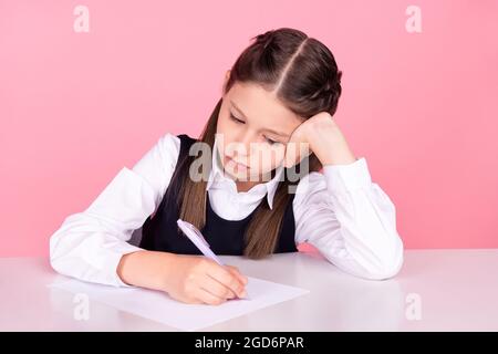 Foto ritratto bambina con code seduta alla scrivania a scuola scrittura test isolato colore rosa pastello sfondo. Foto Stock