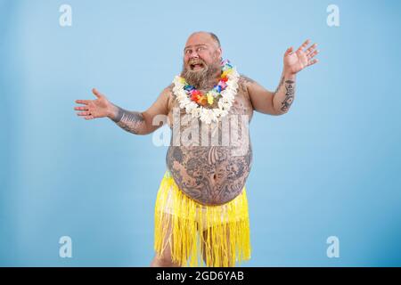 Uomo grasso positivo con tatuaggi in gonna d'erba decorativa danze su sfondo azzurro Foto Stock