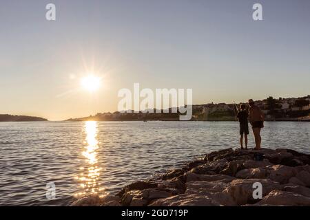 Rogoznica, Croazia-7 luglio 2021: Nonno insegna a suo nipote trucchi di pesca sulla costa rocciosa di Rogoznica, Croazia al tramonto Foto Stock