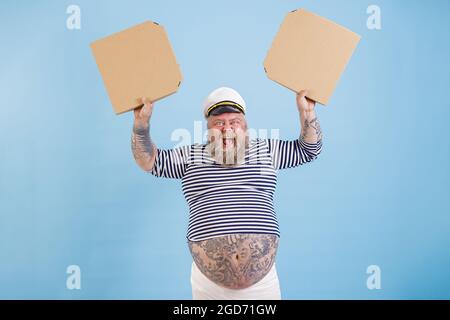 L'uomo gioioso con sovrappeso in costume marinaio tiene scatole vuote di pizza su sfondo azzurro chiaro Foto Stock