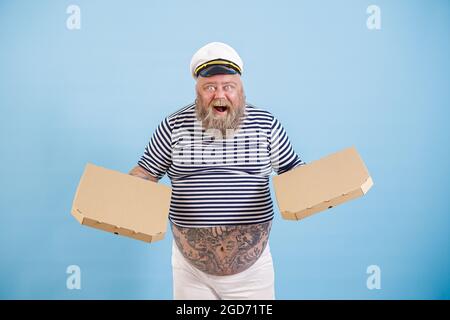 L'uomo eccitato con il peso eccessivo in costume marinaio tiene scatole di pizza su sfondo azzurro chiaro Foto Stock