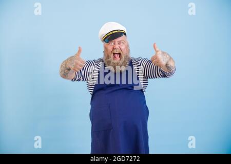 La persona maschile emozionale con sovrappeso nella tuta marinara a mostra il pollice su sfondo azzurro Foto Stock