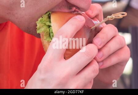 Un uomo mangia un grande hamburger succoso in un negozio di hamburger. Hamburger con formaggio, verdure, cutlets primo piano. Foto Stock