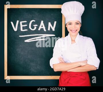 Donna chef menu vegan con uniforme che mostra il cartello nero al ristorante per lo speciale di oggi. Cibo vegeteriano. Parola VEGAN scritto a mano su nero Foto Stock