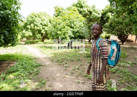 Felice piccolo studente africano sulla strada per la scuola camminando su un piccolo sentiero sterrato nel mezzo di una foresta di mango Foto Stock