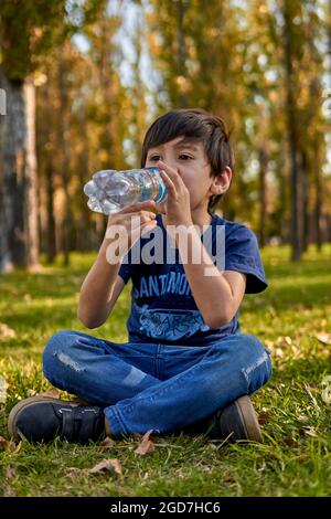 bambino latino che beve acqua da una piccola bottiglia seduta nel mezzo della foresta in autunno. sfondo verticale, sfocato Foto Stock