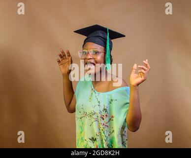 Una donna o una laureanda nigeriana africana, indossando un cappello e bicchieri di laurea, che esprime gioia per il successo nella sua scuola, istruzione e carriera Foto Stock