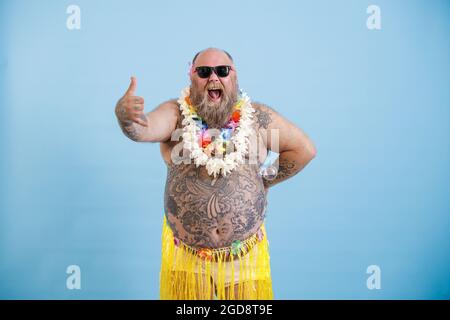 L'uomo allegro con occhiali da sole in gonna d'erba mostra il gesto di saluto Shaka su sfondo blu Foto Stock