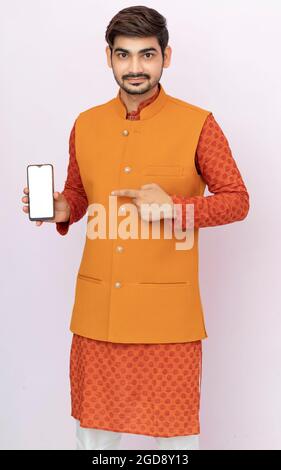 Ritratto di un giovane allegro che indossa etnico indiano tradizionale abbigliamento sfondo isolato, mostrando schermo vuoto telefono cellulare. Foto Stock