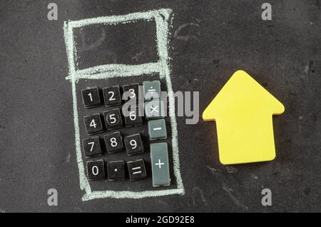 Finanza, concetto immobiliare. Una casa di carta gialla e una calcolatrice disegnata su una lavagna. Forma calcolatrice disegnata a mano con bottoni in plastica. Vista dall'alto. Foto Stock