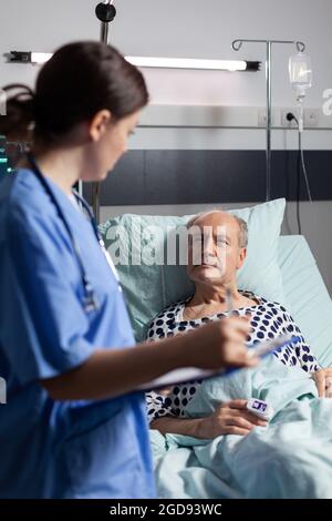 Assistente medico che controlla il trattamento dell'uomo anziano che legge le note sulla clipboard. Ammalato anziano che si trova a letto discutendo con l'infermiere circa l'esperienza di diagnosi con la sacca per infusione attaccata. Foto Stock